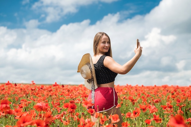 Ukrainian woman in sportswear and straw hat taking a photo selfie with smartphone in poppies field in summer day.