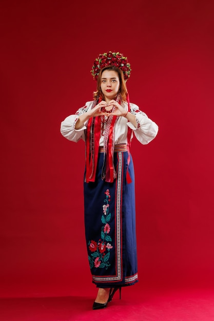 Photo ukrainian woman in traditional ethnic clothing and floral red wreath on viva magenta studio background with hands showing a heart shape ukrainian national embroidered dress call vyshyvanka