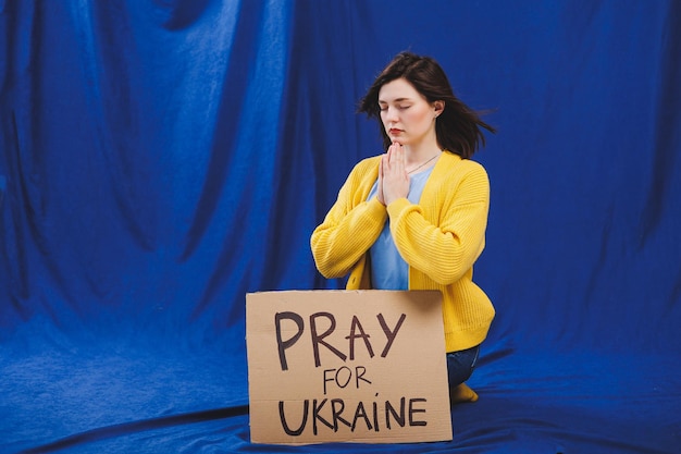 A ukrainian woman in a yellow jacket and blue jeans with a sign\
pray for ukraine ukrainian girl suffered from the war there is no\
war a woman protests against russia\'s aggression against\
ukraine