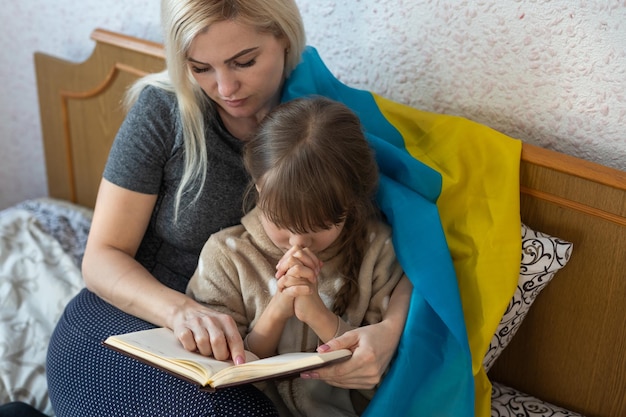 Ukrainian young mother with daughter with flag of ukraine\
praying peace during war conflict between russia and ukraine,\
invasion of russia in ukraine