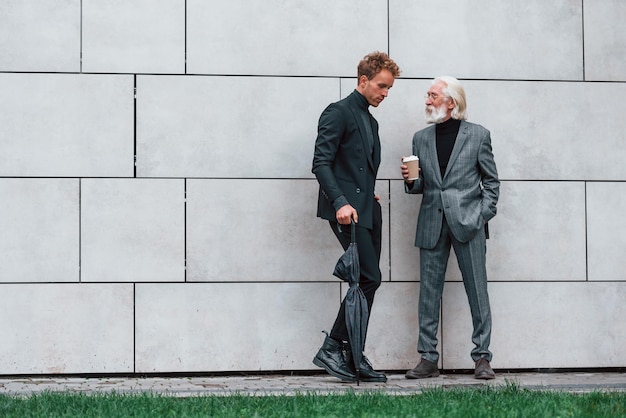 Umbrella and cup of drink Young guy with senior man in elegant clothes is outdoors together Conception of business