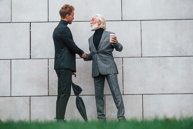 Umbrella and cup of drink Young guy with senior man in elegant clothes is outdoors together Conception of business