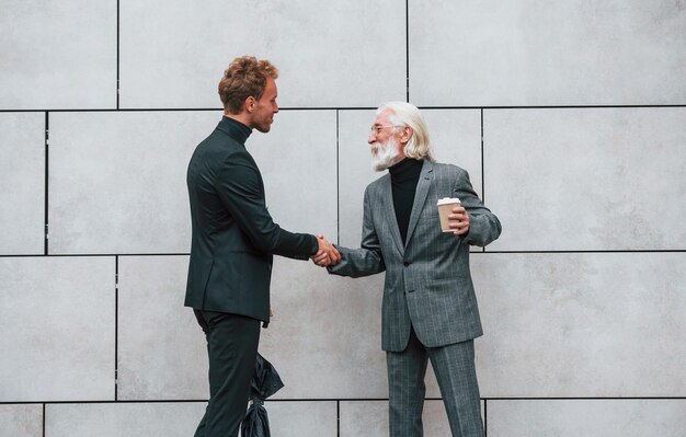 Umbrella and cup of drink Young guy with senior man in elegant clothes is outdoors together Conception of business