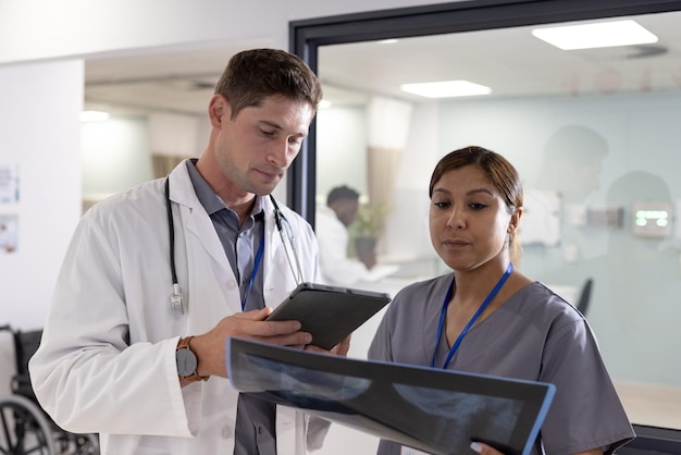 Unaltered portrait of diverse male and female doctor with tablet and x ray in hospital corridor. Hospital, medical and healthcare services.