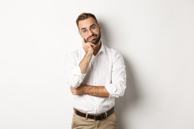 Unamused bearded man in glasses looking displeased at camera, standing moody over white background.