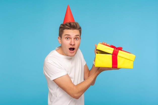 Unbelievable gift Portrait of funny surprised man with party cone on head opening box unpacking present and looking at camera with astonishment indoor studio shot isolated on blue background