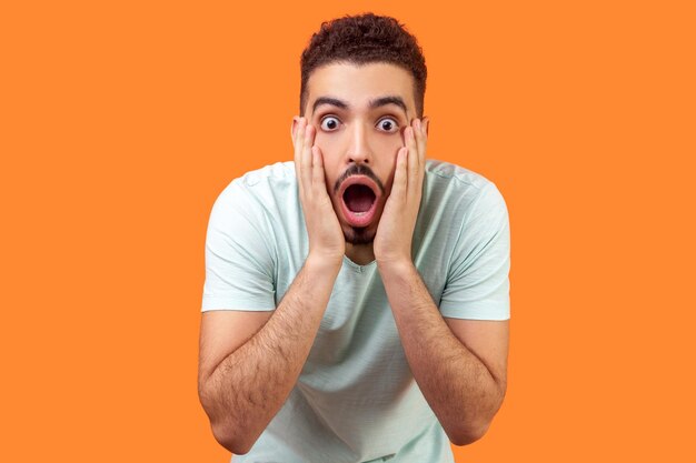 Unbelievable shocking news Portrait of stunned brunette man with beard in casual white tshirt holding face and keeping mouth wide open in amazement indoor studio shot isolated on orange background