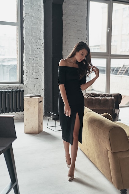 Unbelievably beautiful. Full length of attractive young woman in elegant black dress with a deep slit touching her hair while walking in the room