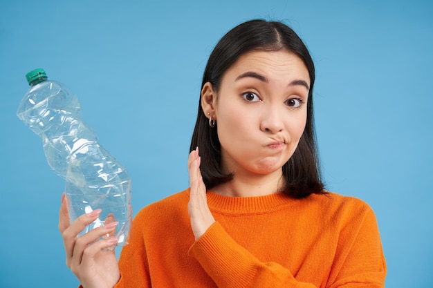 Unbothered asian woman holds plastic bottle and shows block sign refuses to recycle blue background