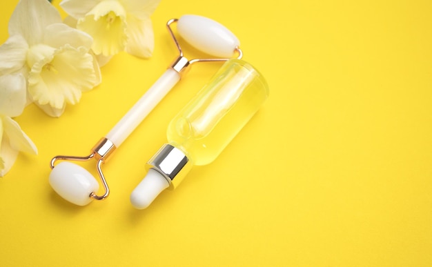 Unbranded serum bottle face roller and narcissus flowers on yellow background Cosmetic facial skin care and spa Natural treatment concept Top view Flat lay