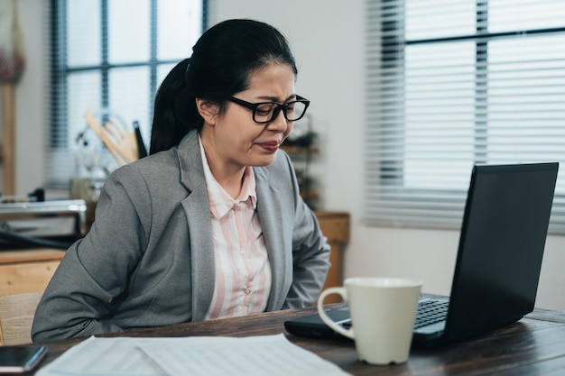 uncomfortable korean female worker pressing her belly is having sudden ache in her stomach while teleworking in apartment. asian lady in pain from menstruation can't continue working on project.