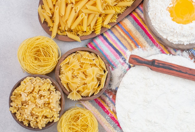 Uncooked different types of pasta with flour and raw egg on a tablecloth.