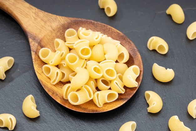 Uncooked elbows macaroni pasta in wooden spoon on dark table.