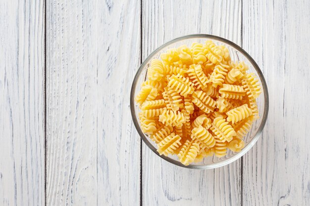 Uncooked radiatori pasta in glass bowl on white wooden background with copyspace