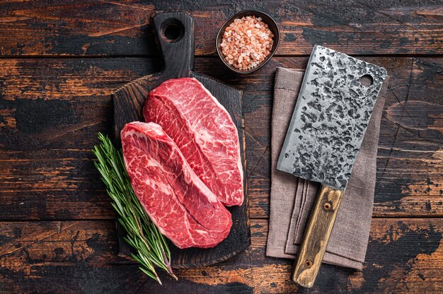 Uncooked raw shoulder top blade or flat iron beef meat steaks\
on a wooden butcher board with meat cleaver. dark wooden table. top\
view.