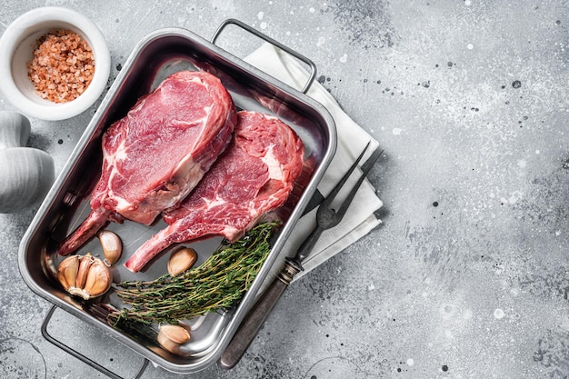 Uncooked Raw Tomahawk veal steaks in steel tray with herbs. Gray background. Top view. Copy space.