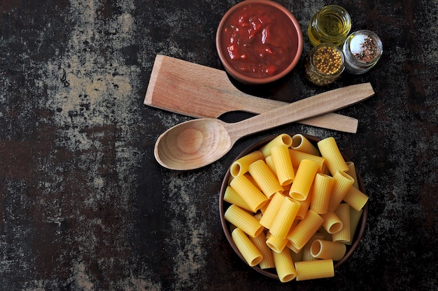 Uncooked rigatoni pasta in a bowl. Tomato sauce and spices. Wooden spatula and spoon. Concept cooking pasta with tomato sauce. Top view. Flat Lay.