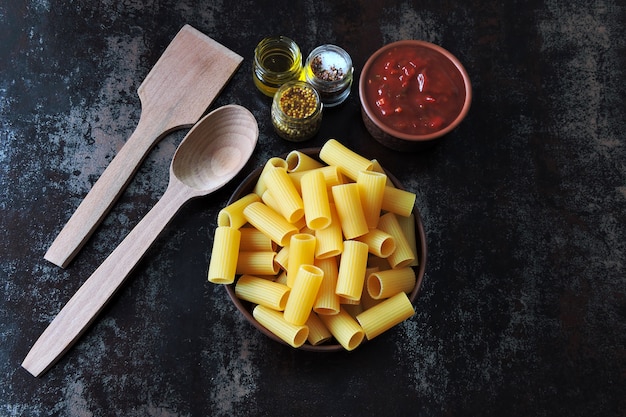 Uncooked rigatoni pasta in a bowl. Tomato sauce and spices. Wooden spatula and spoon. Concept cooking pasta with tomato sauce. Top view. Flat Lay.