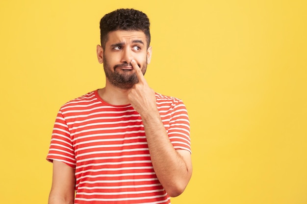 Некультурный скучающий бородатый мужчина в красной полосатой футболке засовывает палец в нос и бурит, дурачится, показывая свои плохие манеры. Крытая студия снята на желтом фоне