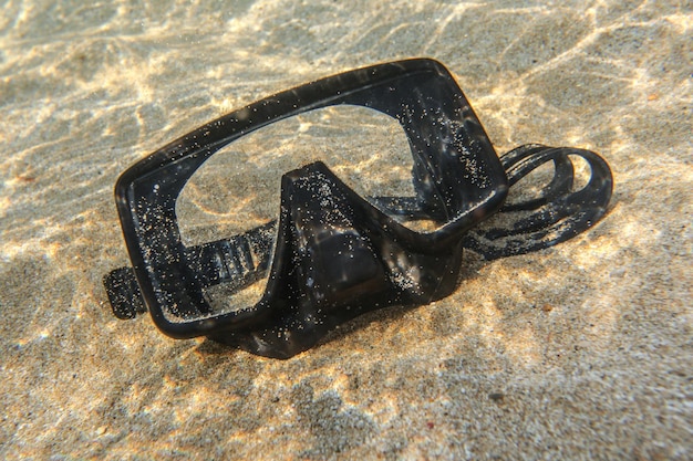 Underwater photo - sun shines on black diving mask on sand in shallow water near beach