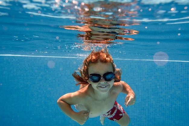 きれいな透明なプールの水に飛び込み、青い背景にカメラを見ている水泳パンツとゴーグルを着た陽気な子供の水中撮影