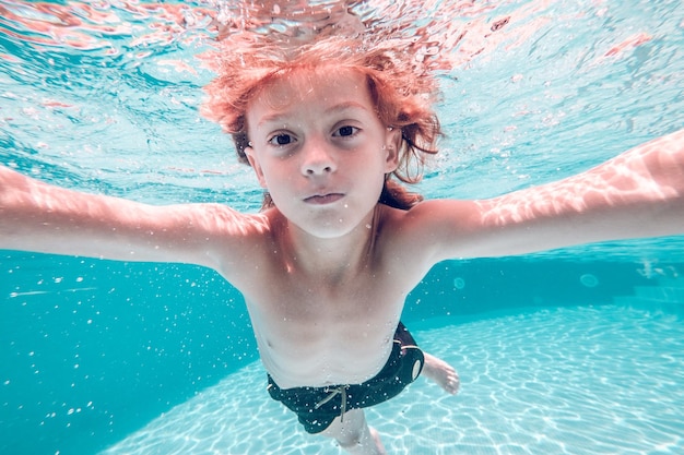 Underwater view of kid with naked torso and in swimming shorts swimming in clean transparent pool water and looking at camera