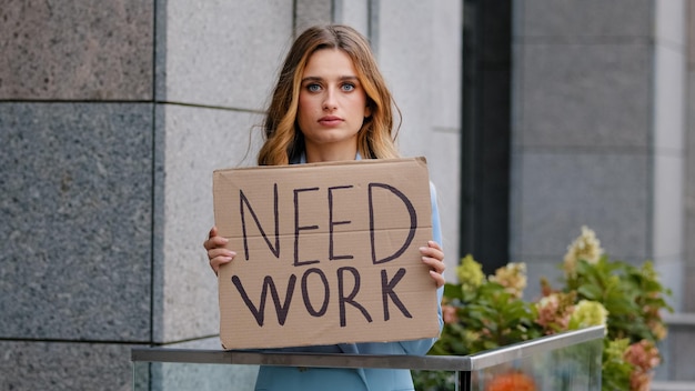 Photo unemployed blonde business woman stands in city looking at camera holds cardboard banner needs work