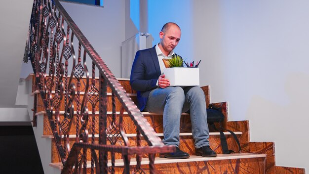 Unemployed businessman sitting on office stairs in depression with box of stuff, lost his business. Group of professional successful businesspeople working in modern financial building.