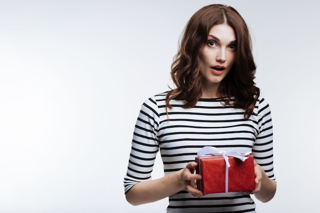 Unexpected surprise. Gorgeous auburn-haired young woman holding a box with a present and looking surprised to receive it