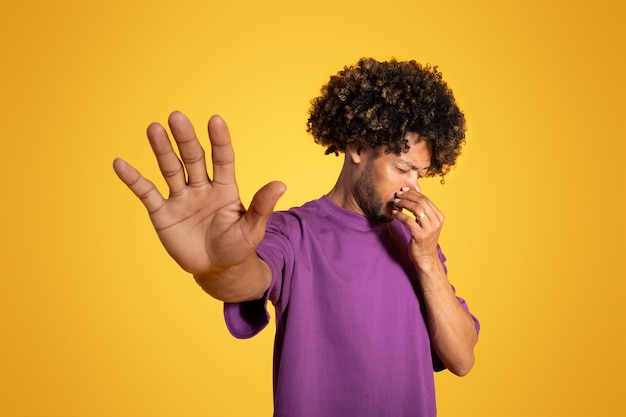 Unhappy adult african american curly man in purple tshirt covering his nose making stop sign