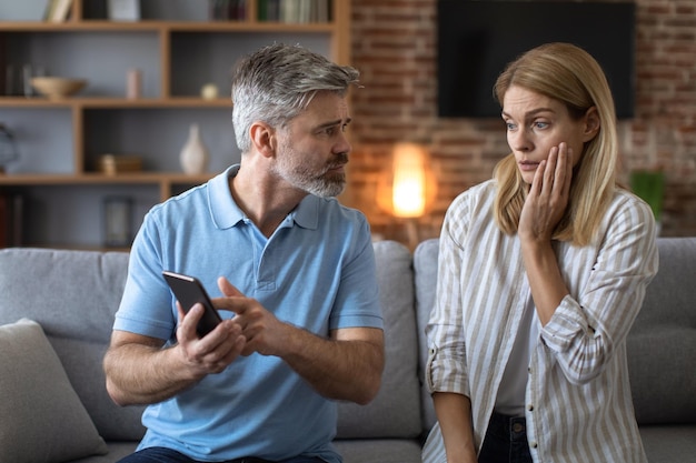unhappy adult caucasian man with beard shows phone to shocked lady woman suffers from gadget addiction