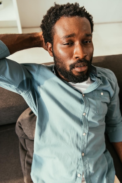 Photo unhappy african american man in sweaty shirt sitting on couch and suffering from summer heat