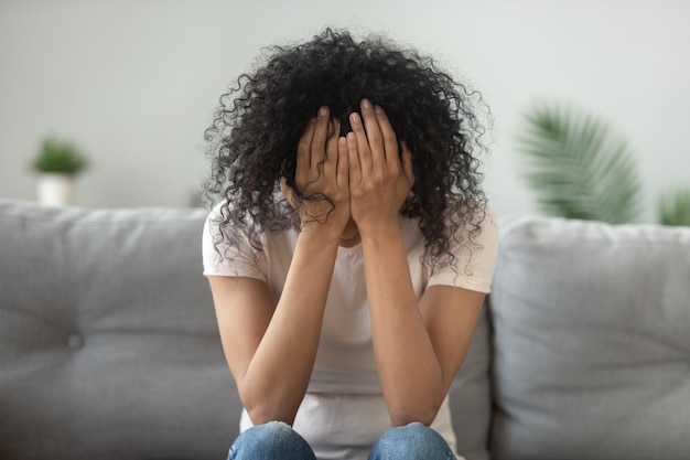 Unhappy African American woman holding head in hands close up crying sitting on couch at home alone upset girl has psychological troubles trauma thinking about problem feeling lonely