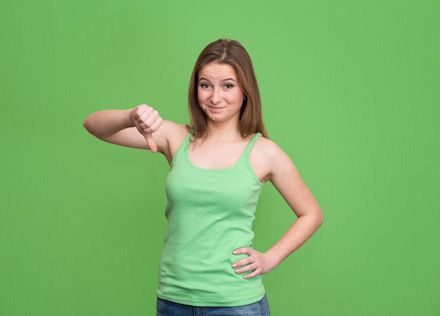 Unhappy angry displeased teenage girl giving thumb down hand gesture isolated on green background