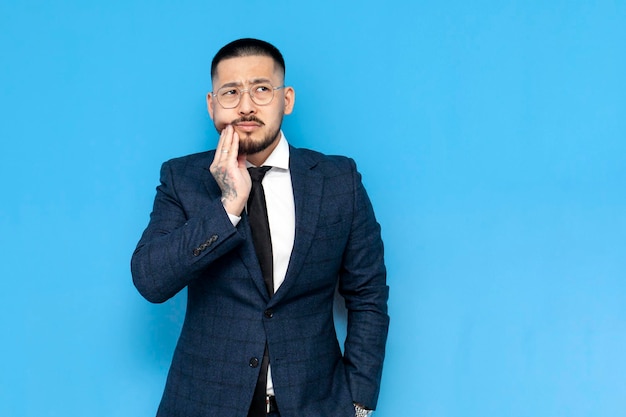 Unhappy asian businessman in suit and glasses holds tooth with his hands on blue isolated background