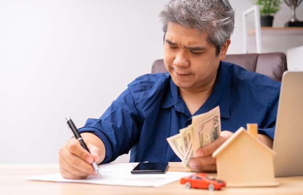 Unhappy Asian man holding a banknote and Making an account of income and expenses to pay the home loan and car loan Concept loan payment and home insurance economic problems and inflation