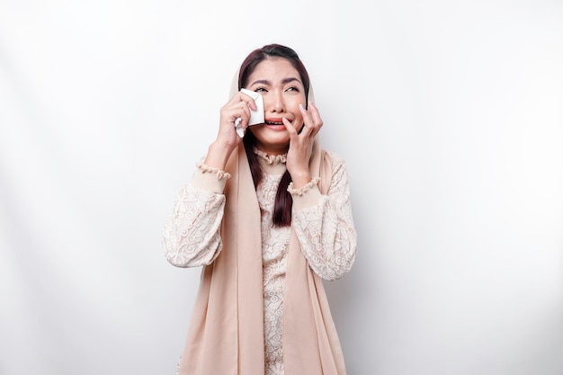 An unhappy Asian Muslim woman wearing a headscarf is wiping her tears in despair feeling depressed and lonely