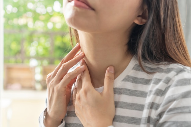 喉の痛み、気分が悪いアジアの不幸な女性。痛みを伴う嚥下、喉の強い痛み、自宅で首に手を触れることに苦しんでいる病気の少女。健康問題の概念。