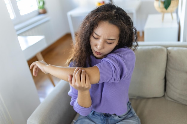 Unhappy asian woman with elbow pain indoors office syndrome health care concept upset frowning asian lady confused looking at arms hurting sitting on couch at home in cozy bright apartment