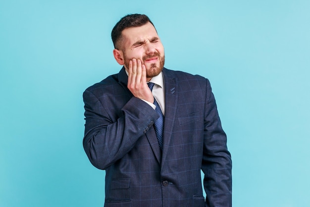 Несчастный бородатый мужчина в официальном костюме чувствует зубную боль, касаясь воспаленной щеки, страдает от кариеса, треснутых зубов, рецессии десны. Снимок в помещении, изолированный на синем фоне