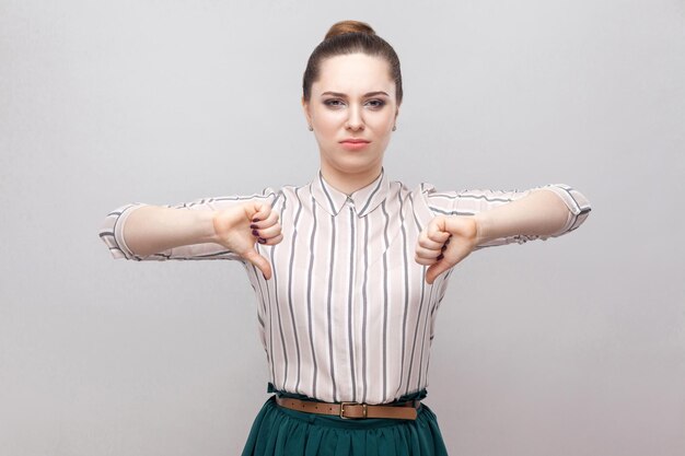 Unhappy beautiful young woman in striped shirt and green skirt with makeup and collected ban hairstyle, standing with thumbs down and looking at camera. indoor studio shot, isolated on grey background