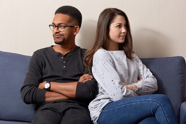 Unhappy couple of different race sit back, not speak to each other after quarrel or dispute, keep hands crossed, misunderstanding and disagreement.