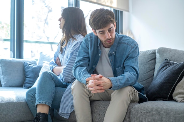unhappy couple sitting on sofa at home looking to opposite directions sad young adult man and woman upset and concerned people with relationship problems to solve living together in apartment