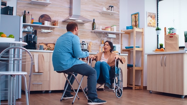 Unhappy disabled wife in wheelchair because of disagreement\
with husband in kitchen. woman with paralysis handicap disability\
handicapped difficulties getting help for mobility from love and\
relations