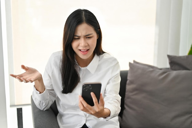 Unhappy and dissatisfied Asian woman sitting on sofa receiving unreasonable news or message