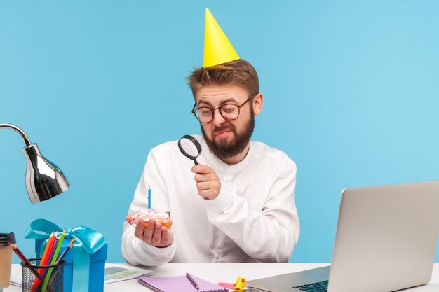 Unhappy dissatisfied man in party cap looking at cupcake with candle through magnifying glass disappointed with little sweet celebrating alone at work Indoor studio shot isolated on blue background