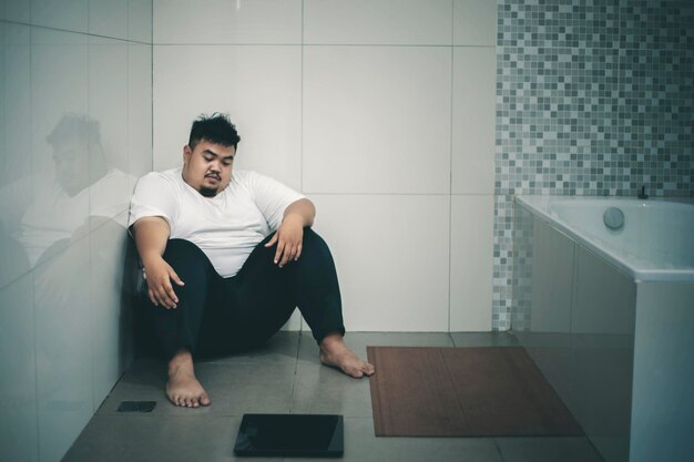 Photo unhappy fat man sits with a scale in the bathroom