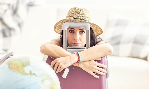 Unhappy female traveler holds suitcase placed next to a globe looking desperate due to travel restrictions.