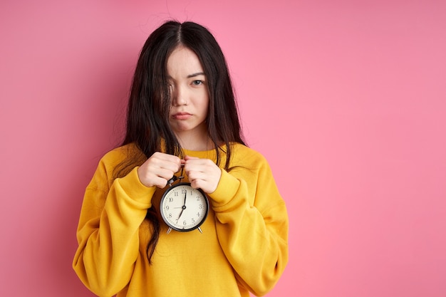 Unhappy female with alarm clock in hands, need some sleep in the morning, looks at camera with sadness. isolated on pink background