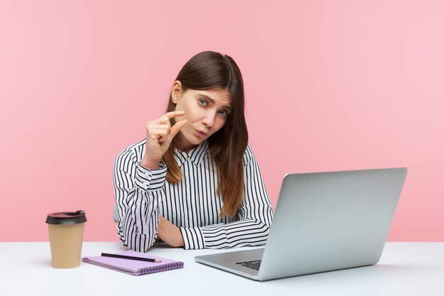 Unhappy frustrated woman office worker sitting at workplace with laptop showing a little bit gesture measuring minimum talking about her salary Indoor studio shot isolated on pink background